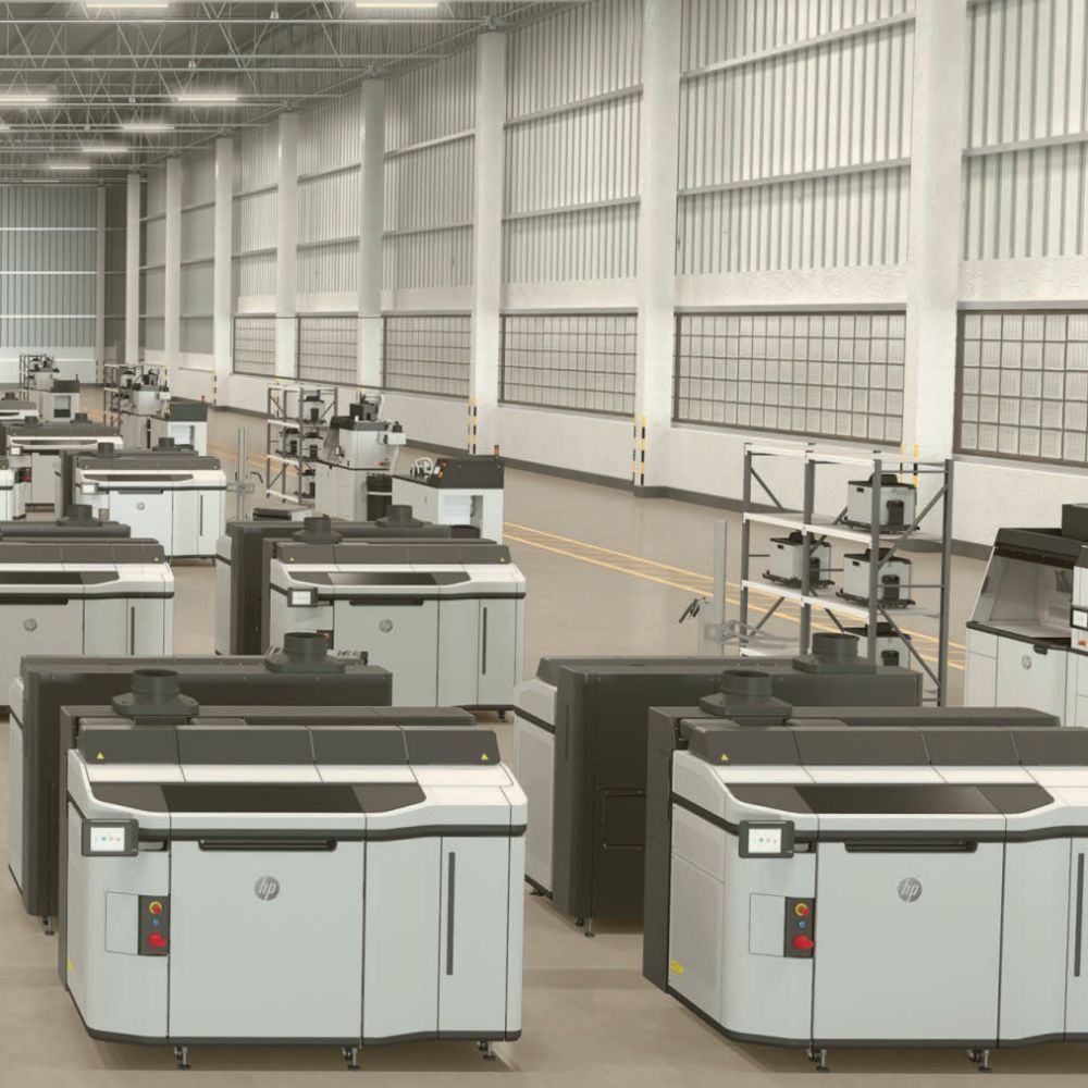 A spacious industrial facility houses multiple large-scale 3D printers, including the advanced HP JF 5200, and other machinery arranged in rows, featuring high ceilings and wide aisles.